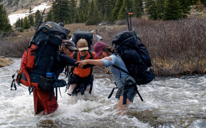 Three people wearing backpacks stand in knee-deep rushing water, bracing themselves with each others' arms. 
